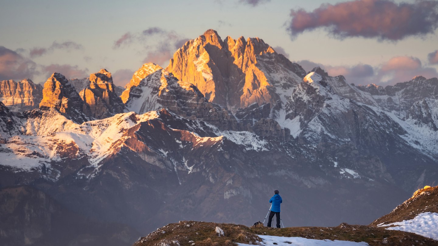 Hiker sets up tripod on snowy ridge overlooking mountains.